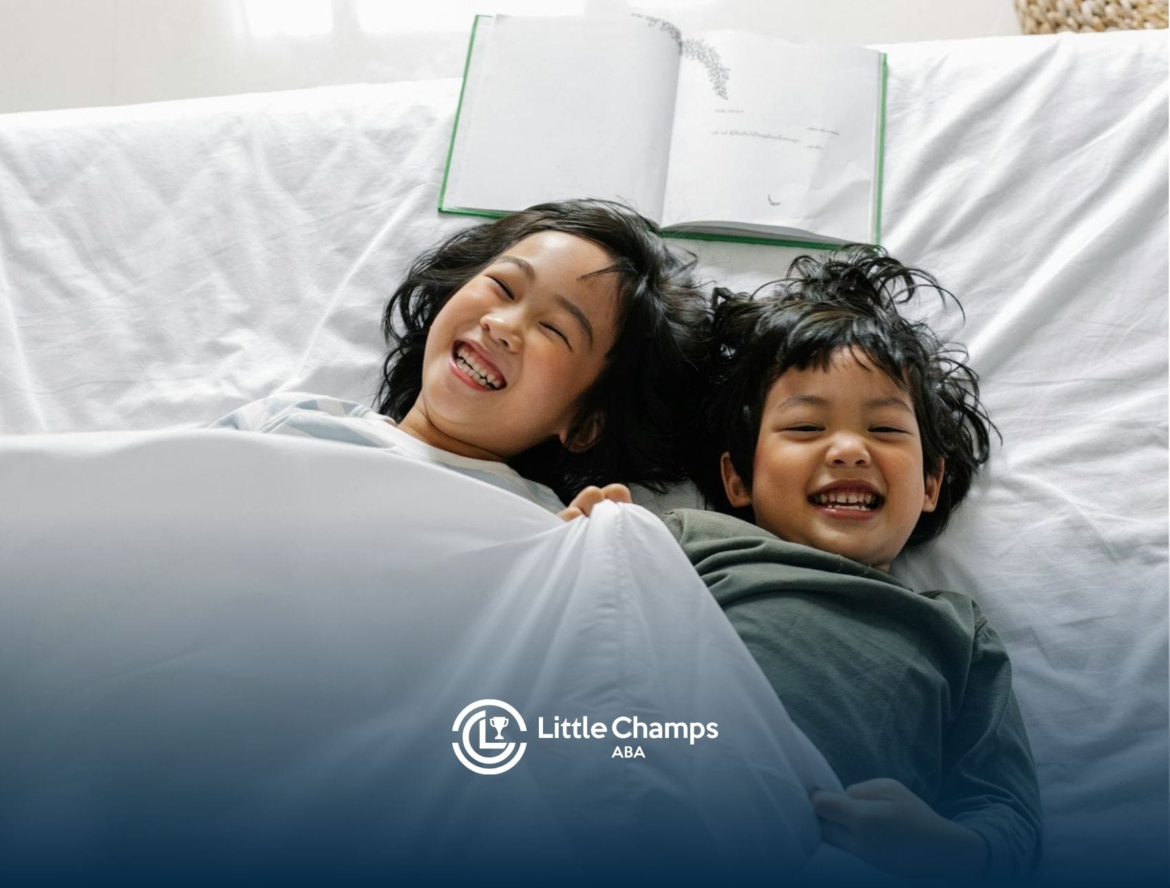 Two smiling autistic children lying in bed under a white blanket with an open book above them in CO.