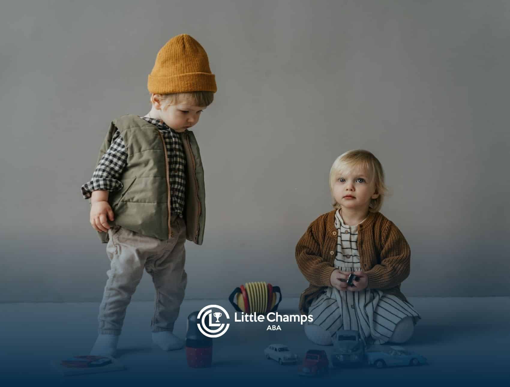 Two autistic toddlers sitting and standing near toy cars and puzzles on a gray floor in Colorado.